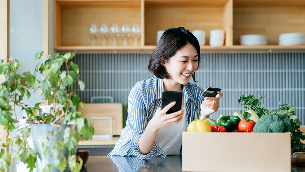 Young woman making a payment on her smartphone with a box of fresh groceries on the kitchen counter at home